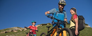 Zwei Mountainbiker schieben ihre Fahrräder vorbei an einer Wanderin. Alle lächeln.