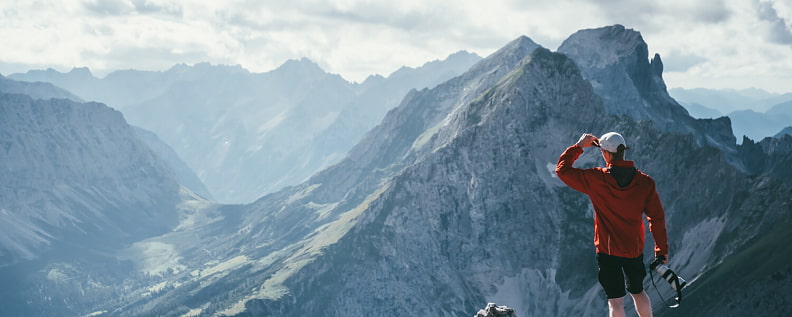 Bergwanderer mit langer roter Bergjacke blickt in die Ferne zu den Bermotiven