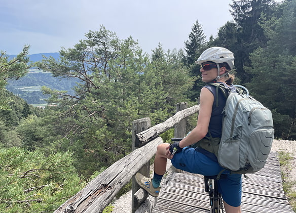 Frau mit Helm und Rucksack sitzt auf einem MTB. Sie steht auf einer Art Holzbrücke und hat ihr Fuß auf dem Geländer abgestellt.