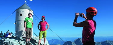 Zwei Menschen stehen am Gipfel, ein Dritter macht mit dem Handy ein Foto.
