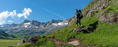 Mann mit Rucksackt steht auf Bergwiese. Im Hintergrund felsige Berge mit etwas Schnee