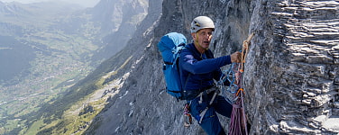 Mann mit Kletterhelm und Rucksack hängt im Klettergurt an einem Stand gesichert an einer Felswand