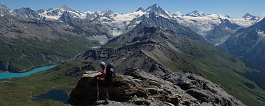 Mensch mit Wanderrucksack steht an einem Berrücken. Dahinter ein See sowie hohe schneebedeckte Berge
