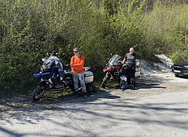 Gerhard und Mathias stehen vor ihren beiden Motorrädern auf einem Parkplatz. Hinter ihnen ist Gebüsch.