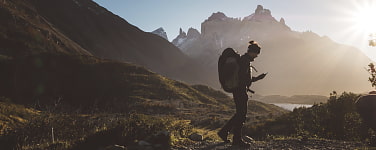 Mann mit Rucksack steht vor einer Bergkulisse. Er blickt auf ein Smartphone in seiner Hand. 